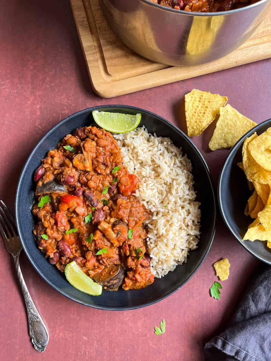 A black bowl of aubergine chilli with rice, lime wedges, a black bowl of tortilla chips, a silver fork and black napkin.