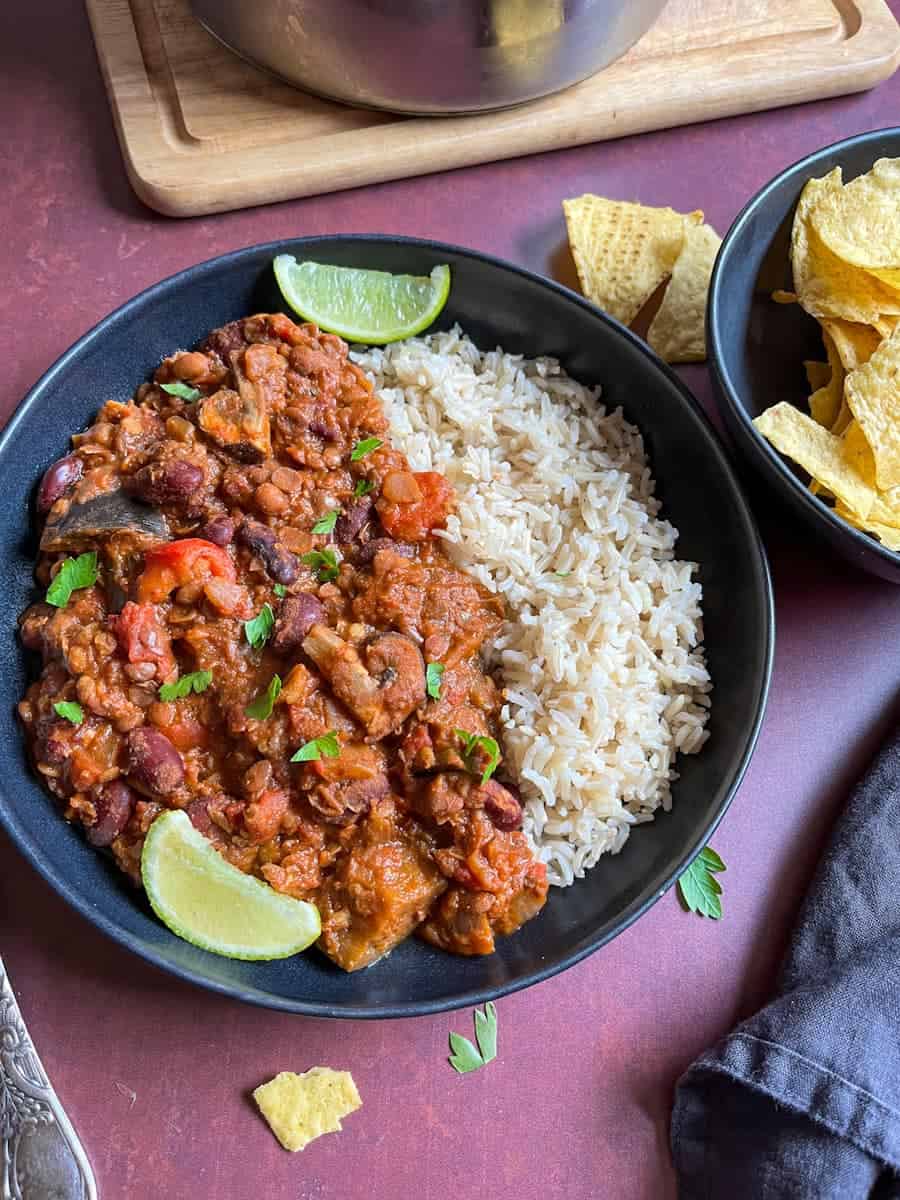 A black bowl of aubergine and lentil chilli with rice and lime wedges and a bowl of tortilla chips.