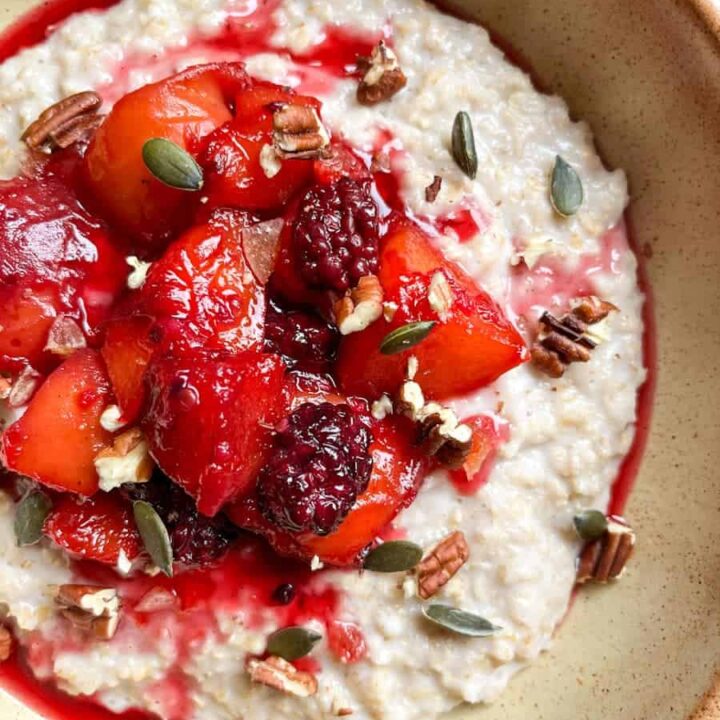 a beige bowl of porridge topped with peach and blackberry compote, pumpkin seeds and pecan nuts.