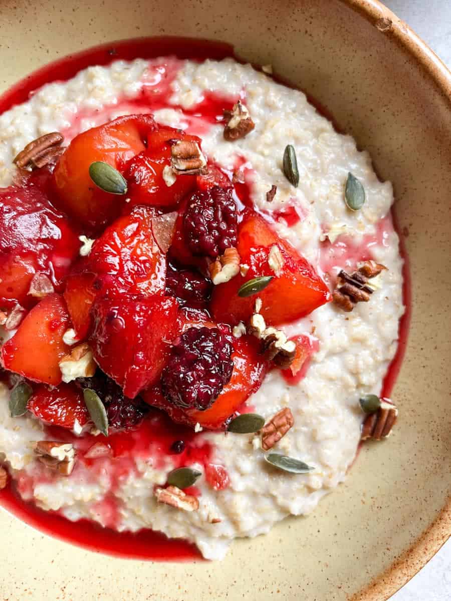 a beige bowl of porridge topped with peach and blackberry compote, pumpkin seeds and pecan nuts.