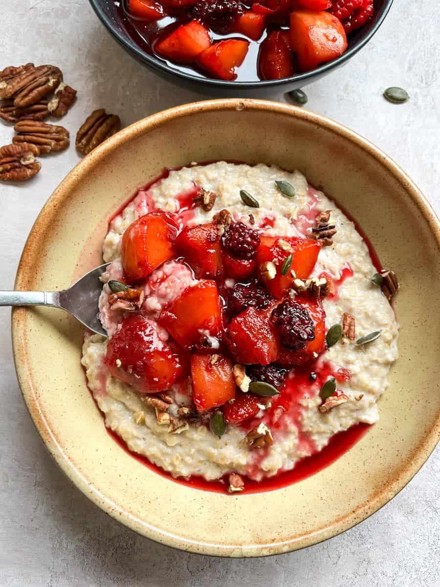 A beige bowl of porridge topped with cooked peaches and blackberries and sprinkled with pumpkin seeds and chopped pecan nuts.