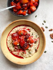 A beige stoneware bowl of porridge with blackberry and peach compote, pumpkin seeds and chopped pecan nuts and a bowl of fruit compote with a silver spoon.