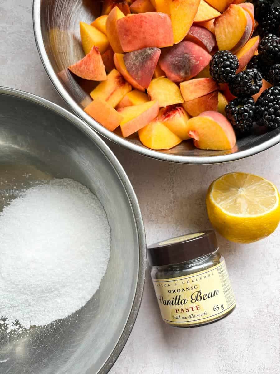 A bowl of white sugar, bowl of sliced peaches and blackberries, half a lemon and a bottle of vanilla bean paste.