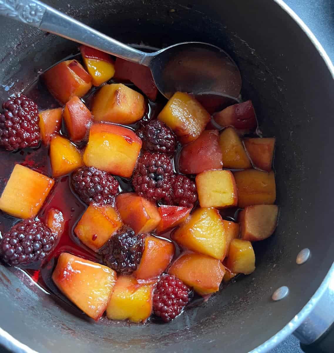 A saucepan with chopped peaches, blackberries and a silver spoon.