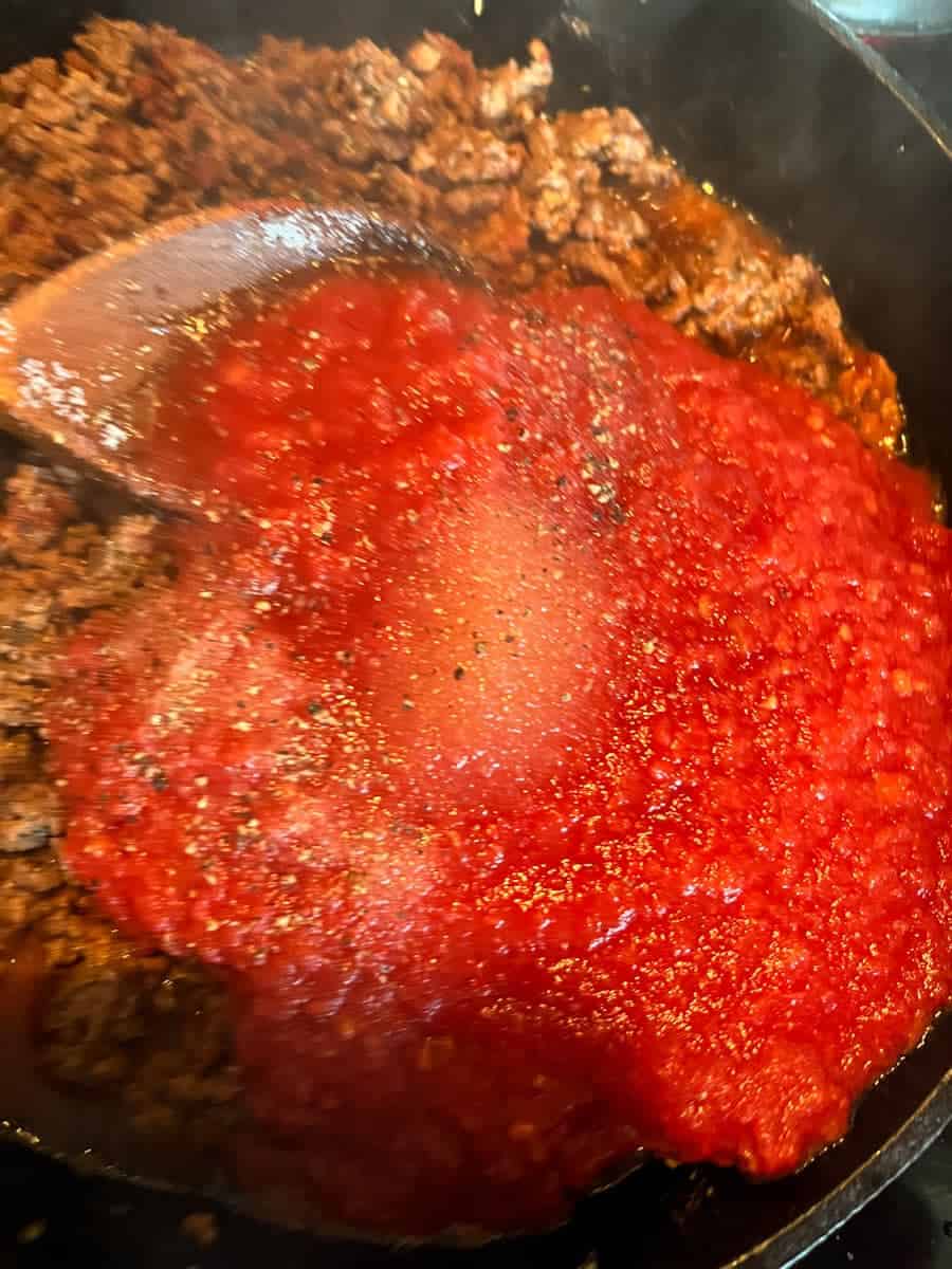 Tomato sauce being stirred into a pan of browned minced beef.