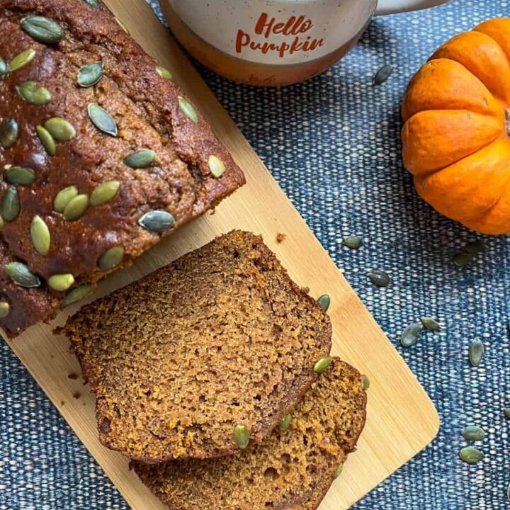 A partially sliced pumpkin loaf cake sprinkled with pumpkin seeds on a wooden serving board, an orange and white mug and a mini pumpkin.