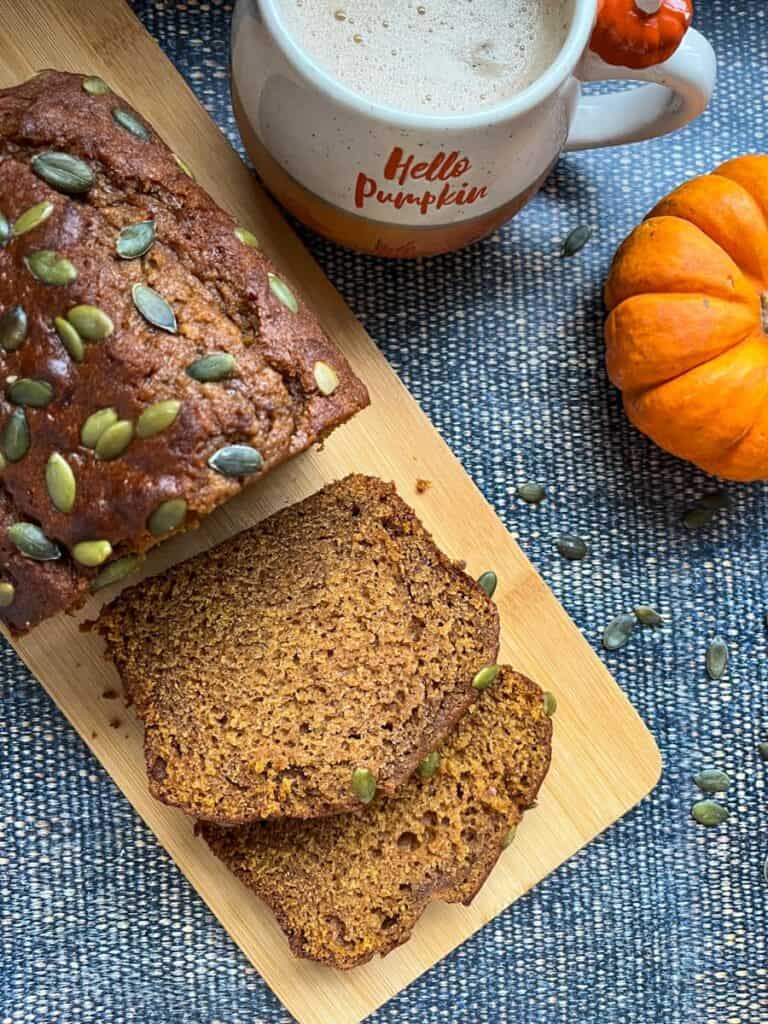 A partially sliced pumpkin loaf cake sprinkled with pumpkin seeds on a wooden serving board, an orange and white mug and a mini pumpkin.