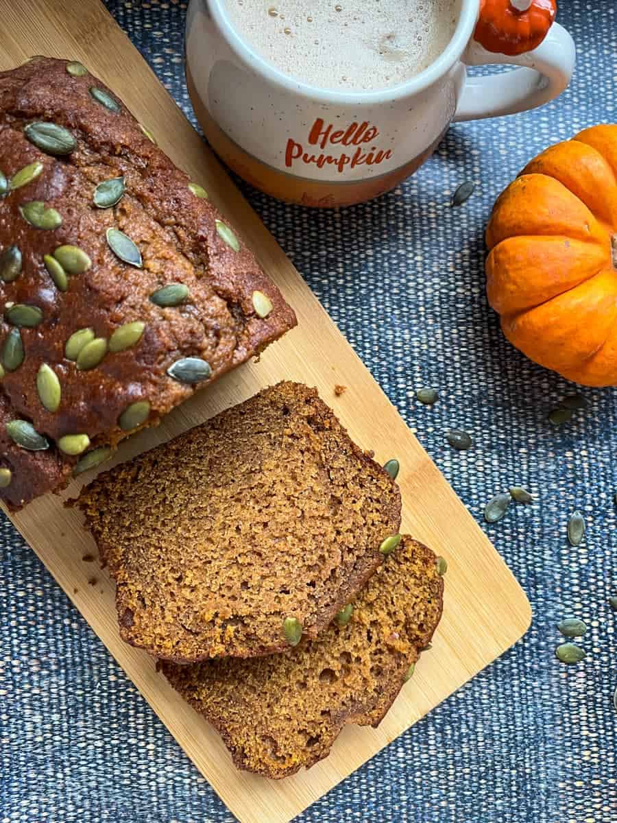 A partially sliced pumpkin bread scattered with pumpkin seeds on a wooden board, an orange and white mug and a mini pumpkin. 