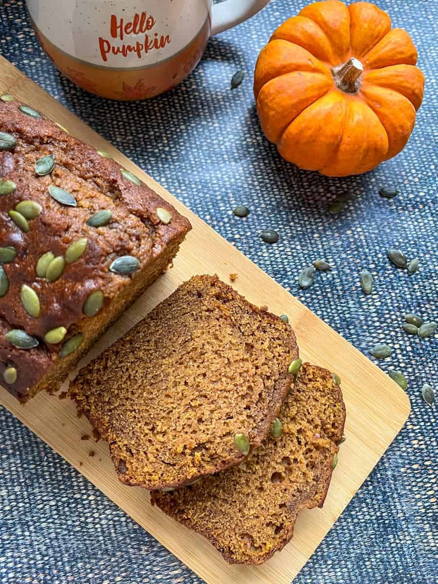 A pumpkin bread topped with pumpkin seeds on a wooden board, an orange and white mug and a small pumpkin.