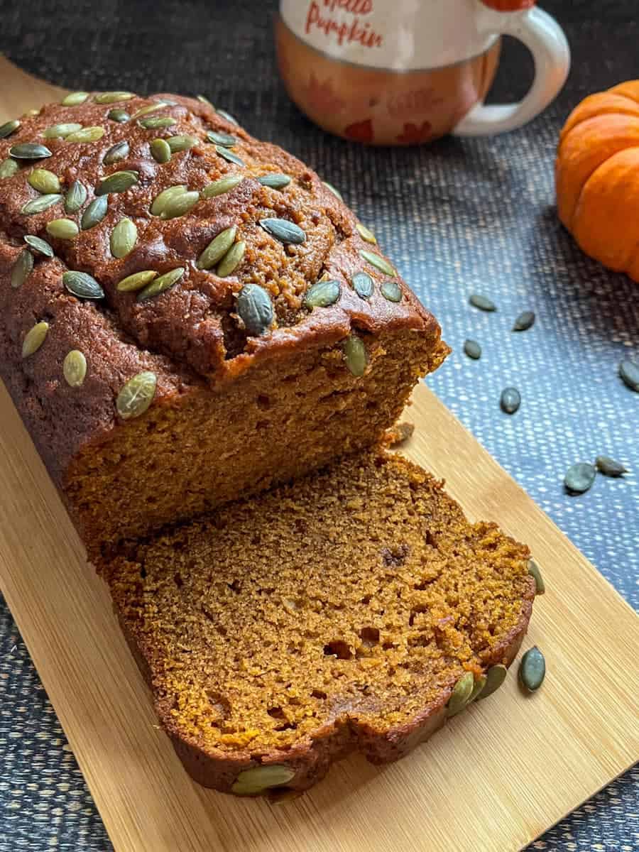 Pumpkin bread with a slice cut from it on a wooden serving board and an orange and white coffee mug.