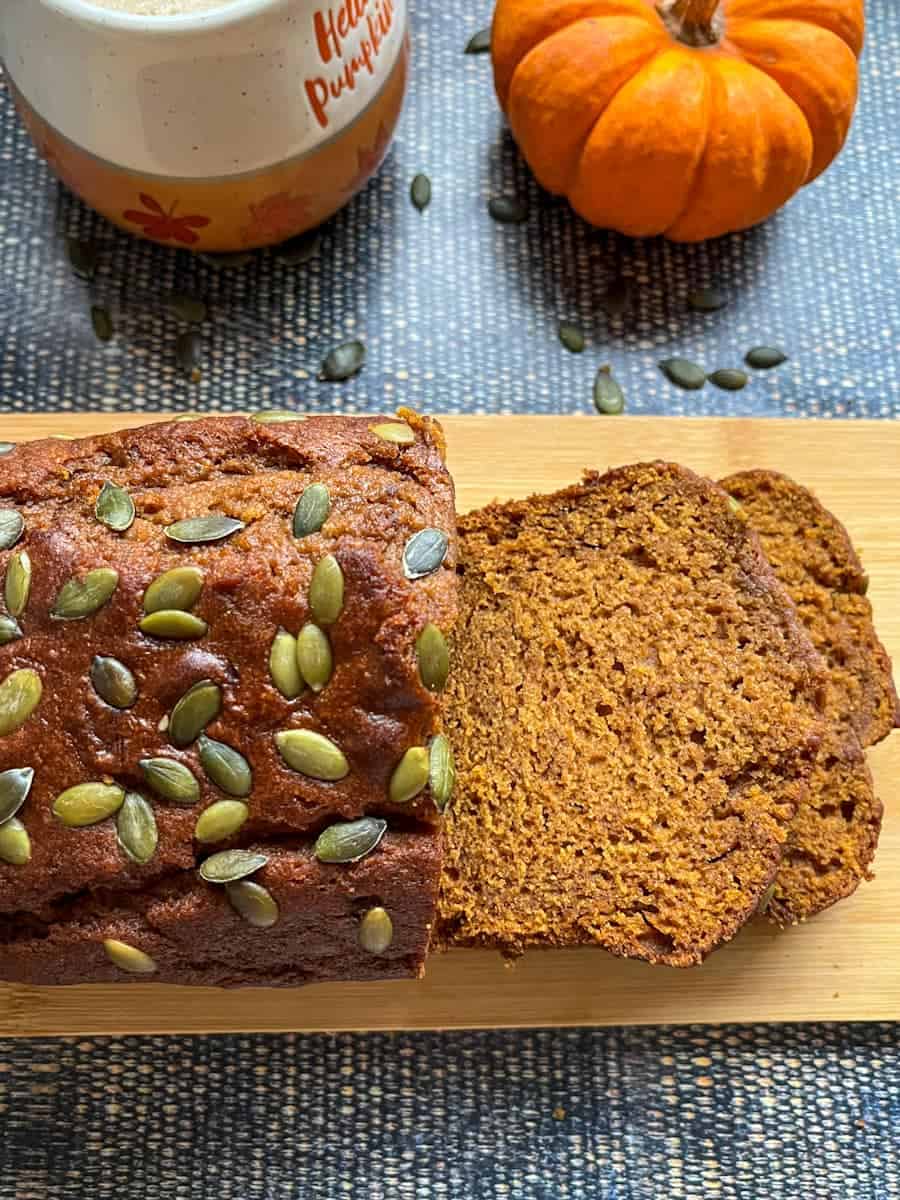 A partially sliced pumpkin loaf sprinkled with pumpkin seeds on a wooden board, an orange and white mug and a munchkin pumpkin.