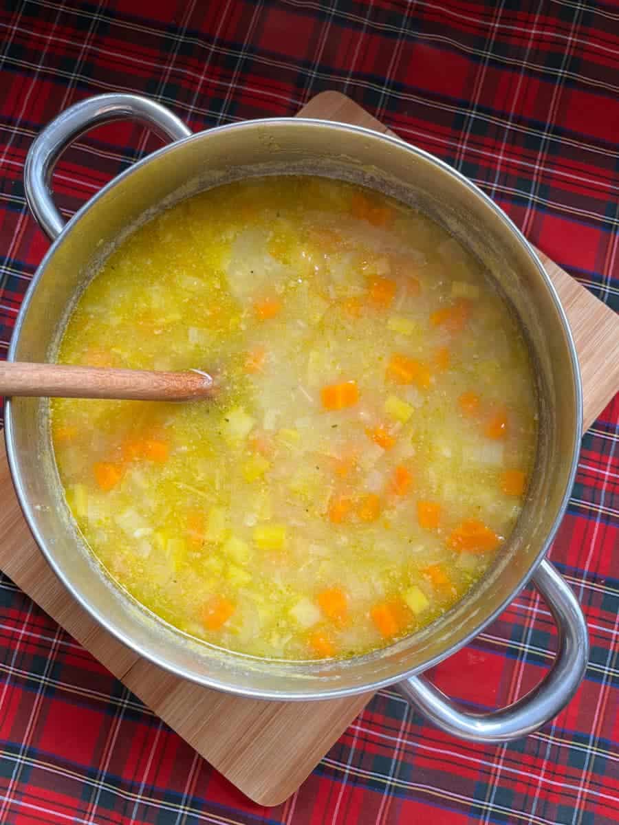 A large silver pan of tattie soup with a wooden spoon on a wooden board and a red tartan tablecloth.
