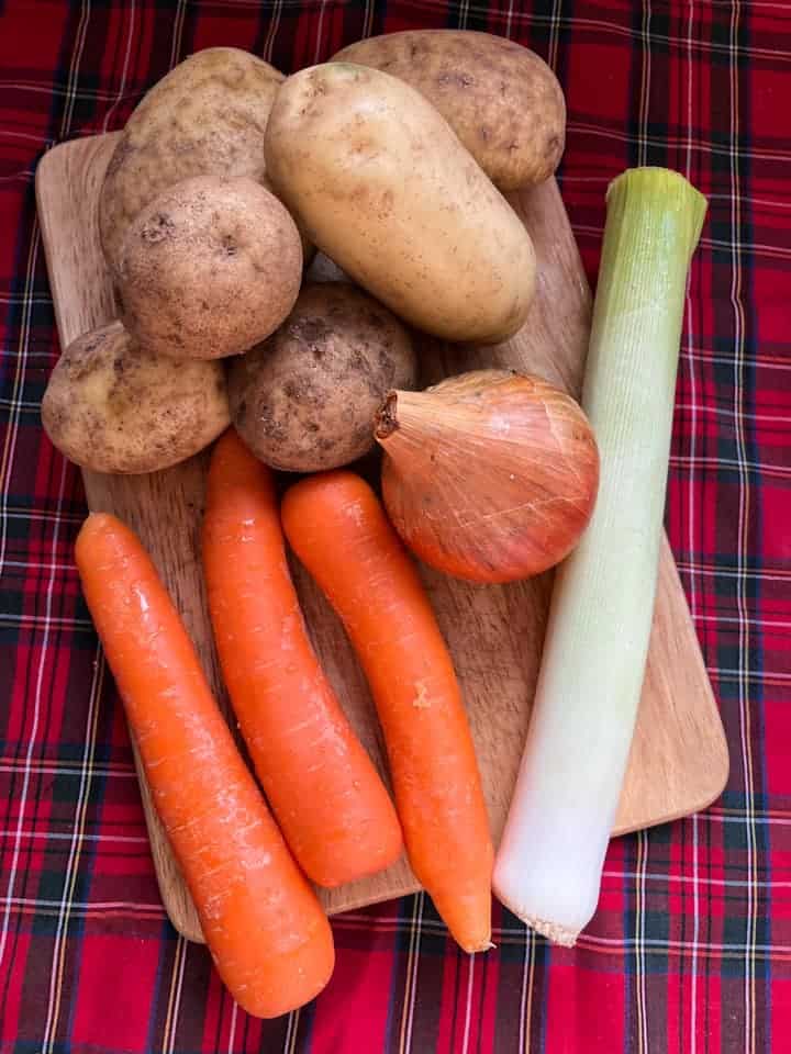 Potatoes, an onion, a leek and three carrots on a wooden chopping board and a red tartan tablecloth.