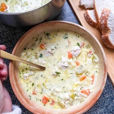 A bowl of creamy chicken soup with rice, diced carrots and leeks with a gold spoon, a wooden board with slices of bread and a large silver pot of soup.