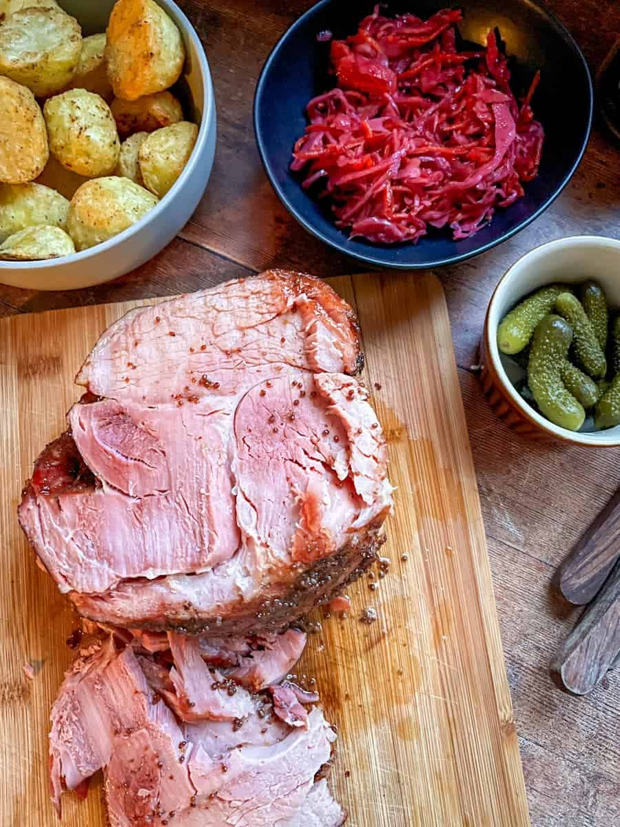 A partially sliced piece of cooked ham on a wooden chopping board, a bowl of potatoes, a black bowl of pickled red cabbage and a small jar of Gherkins.