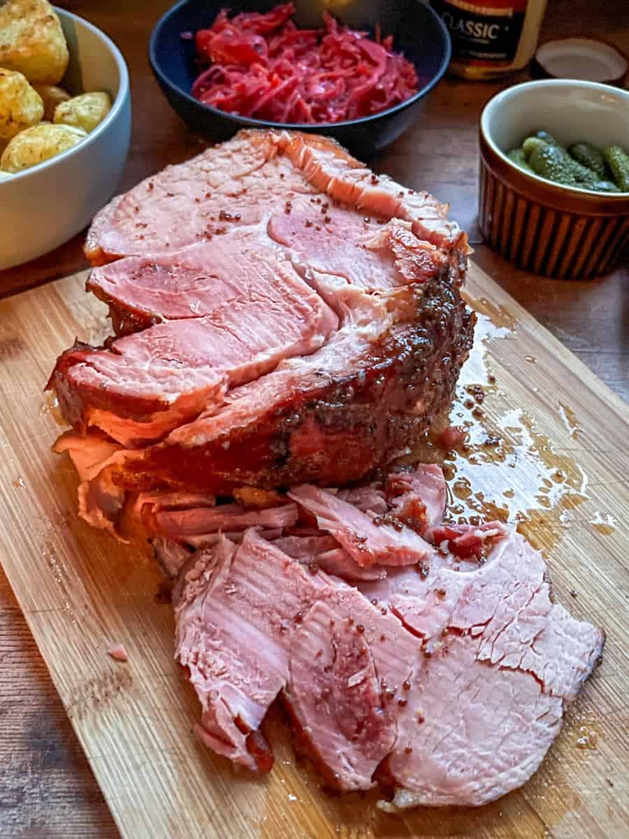A partially sliced cooked gammon joint on a wooden chopping board, a bowl of potatoes, a bowl of pickled red cabbage and a brown pot of Gherkins.