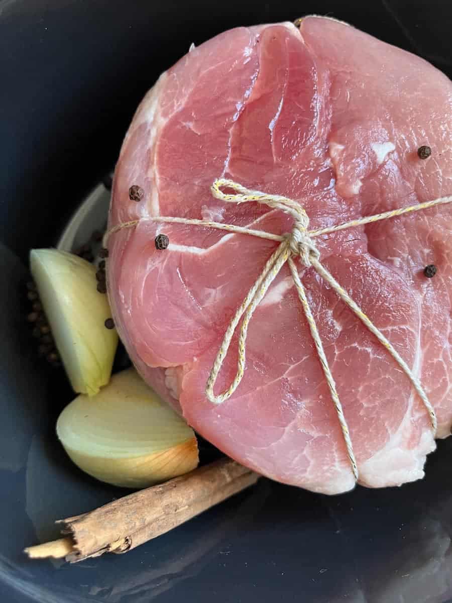 A gammon joint tied up with a piece of string in a slow cooker bowl with a cinnamon stick, black peppercorns and a halved onion.