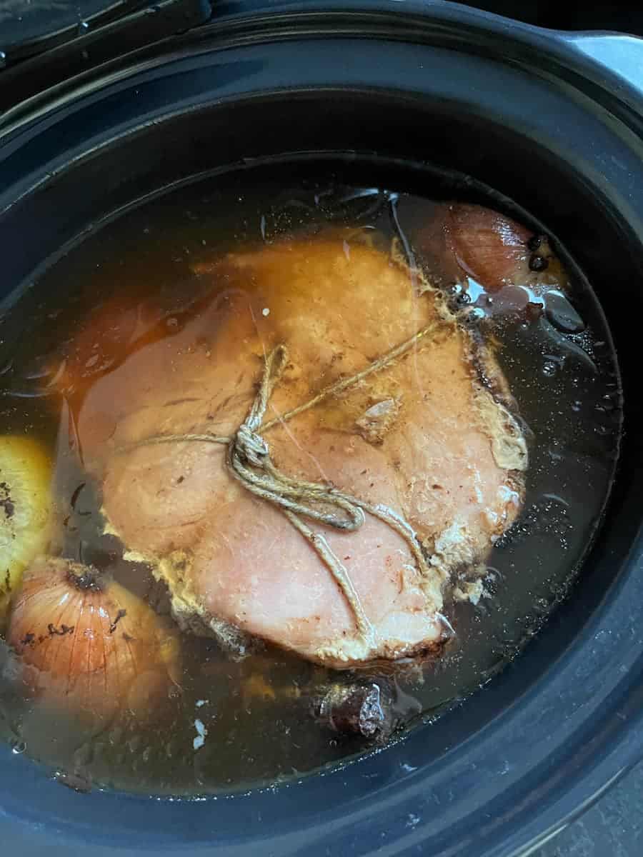 A gammon joint tied with string in a slow cooker with coca cola and onion halves.