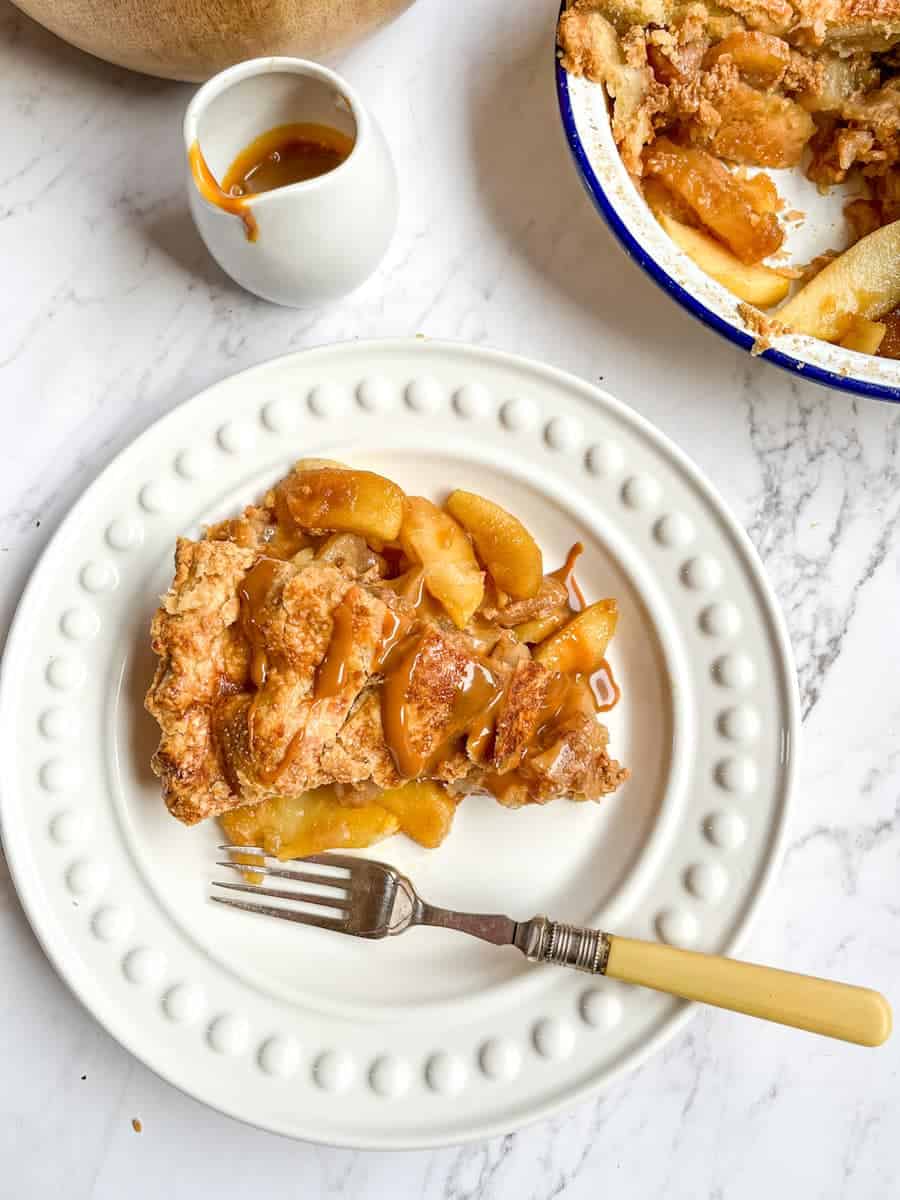A slice of apple pie drizzled with caramel sauce on a white plate with a fork, a jug of caramel sauce and a blue and white dish of the apple pie.