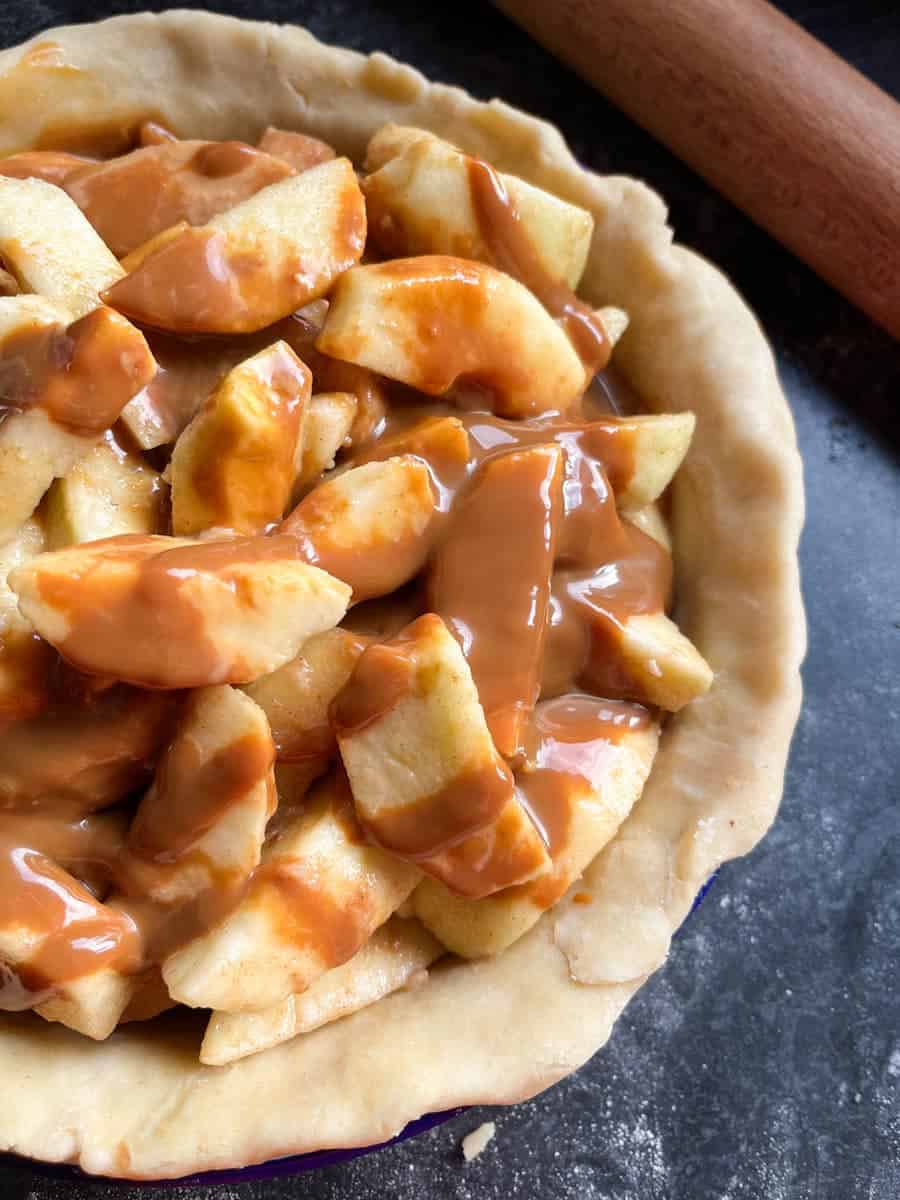 Sliced apples in a pastry lined pie dish drizzled with caramel sauce and a rolling pin.