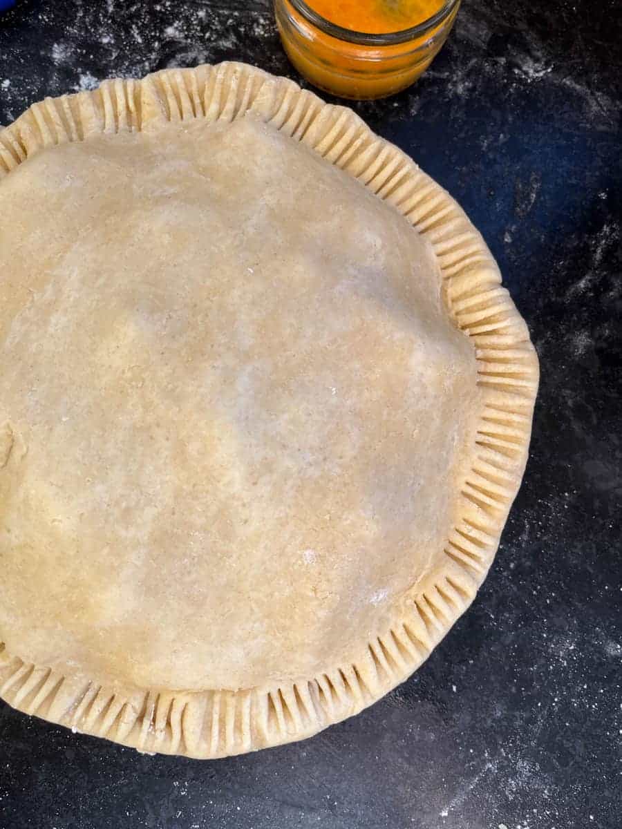 An unbaked apple pie with crimped edges and a jar of beaten egg.