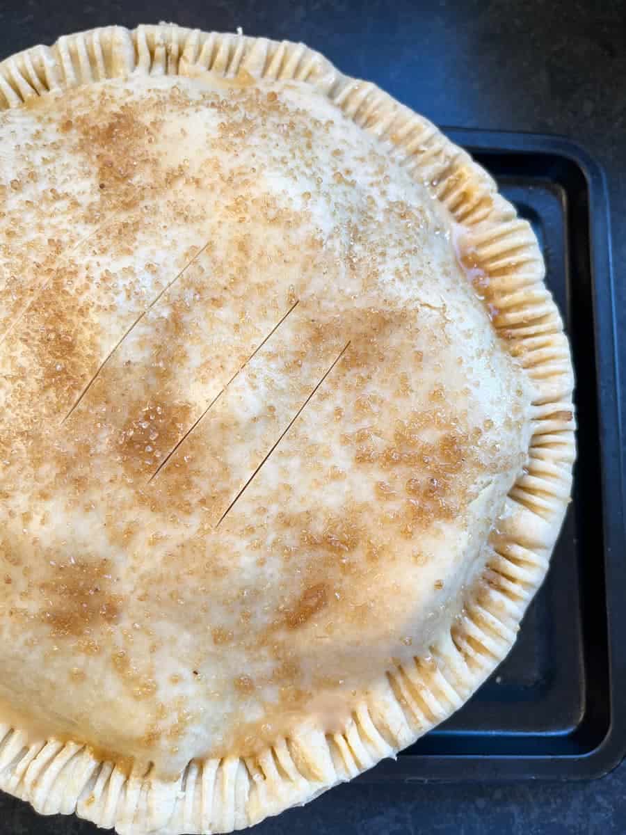 An unbaked apple pie with crimped edges and sprinkled with brown sugar on a black baking tin.