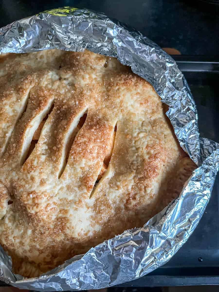 A baked apple pie sprinkled with sugar. A piece of tin foil has been placed over the edge of the pie to prevent the crust from burning.