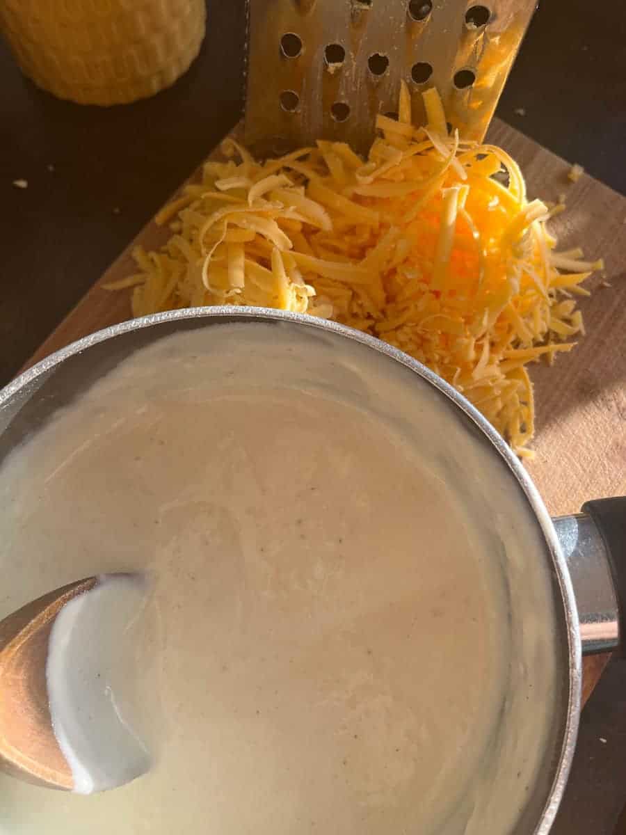 A pan of cheese sauce with a wooden spoon and grated cheese on a wooden chopping board.