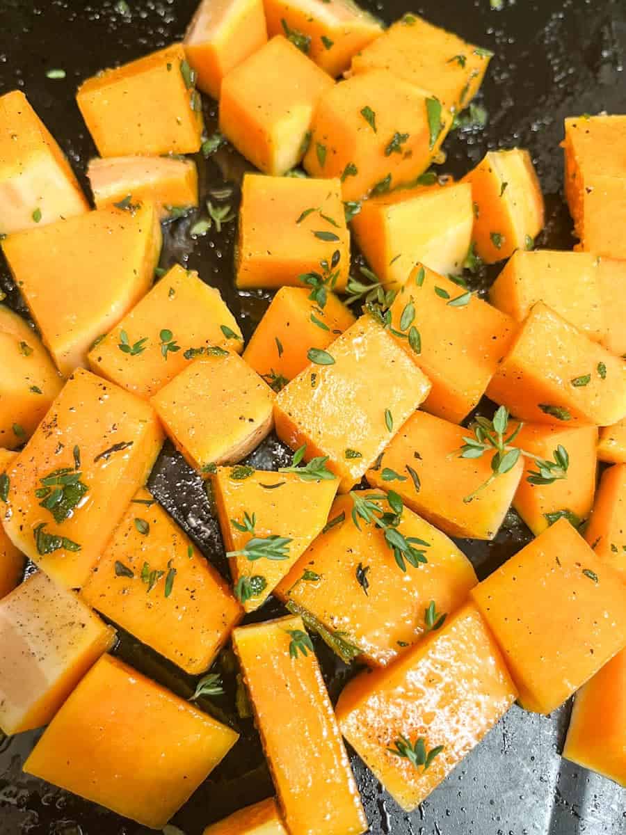 Diced pumpkin covered in fresh thyme and sage on a black baking tin.