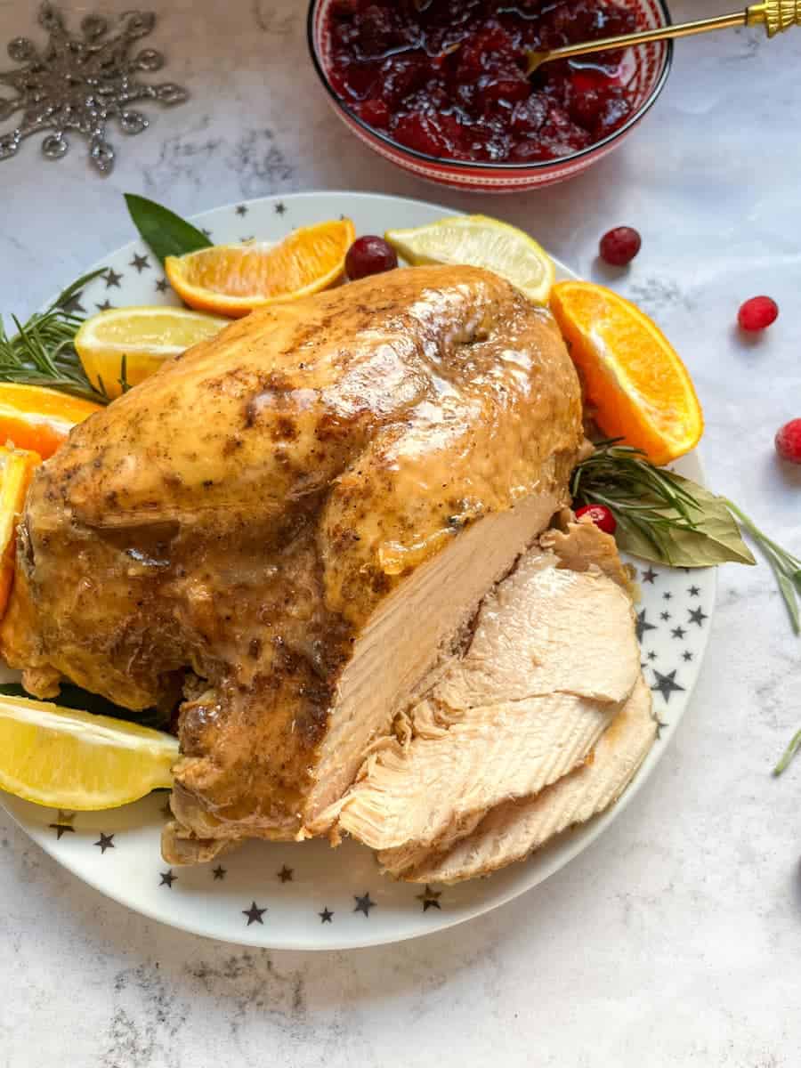 A partially sliced turkey crown on a gold and white star plate with orange and lemon slices and fresh herbs and a bowl of cranberry sauce with a gold spoon.