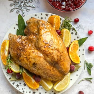 A cooked turkey crown with orange and lemon slices, fresh cranberries and herbs on a white and gold star plate and a bowl of cranberry sauce.