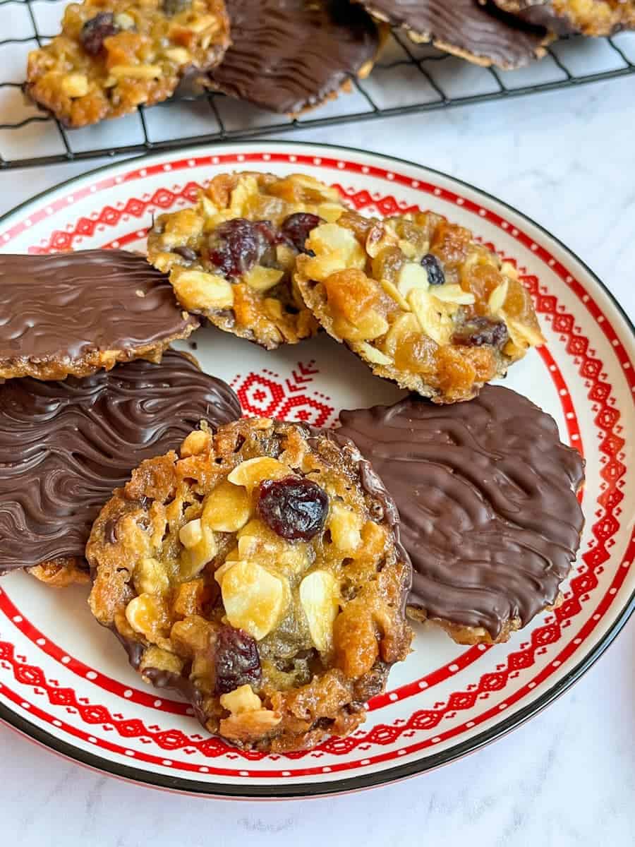 Six Florentines covered in melted dark chocolate on a red and white Christmas plate. More Florentines sit on a wire cooling rack in the background.