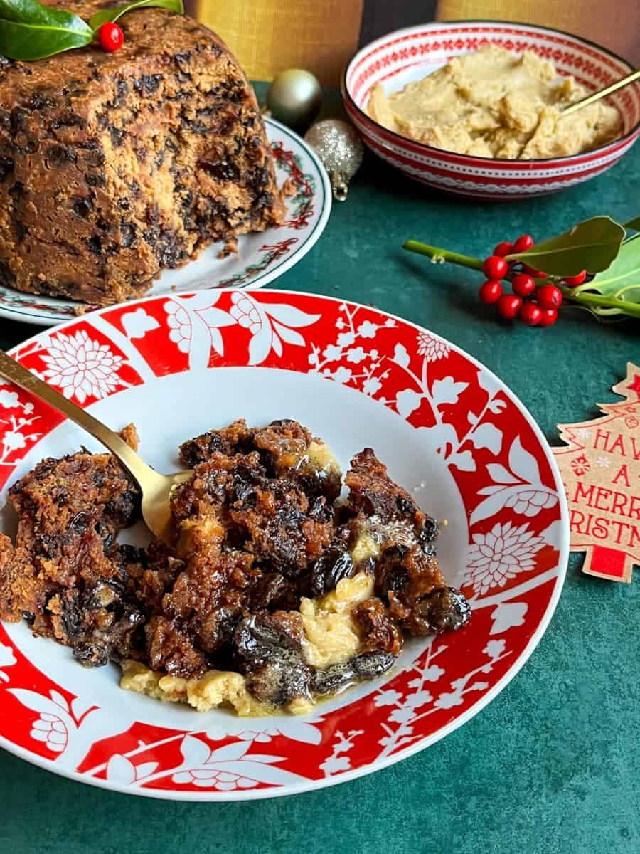 A red and white bowl of Christmas pudding with brandy butter. A Christmas pudding on a festive plate, a red bowl of brandy butter and a sprig of holly sits in the background.