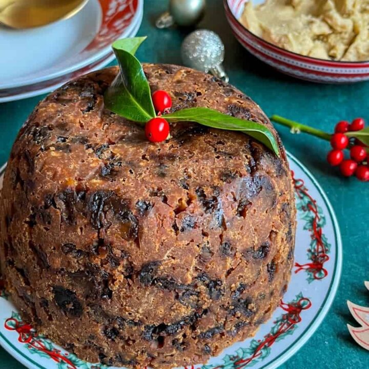 A Christmas pudding on a green and red Christmas plate topped with a sprig of holly, two red and white bowls with gold spoons, a red and white bowl of brandy butter and holly sprigs and Christmas decorations.