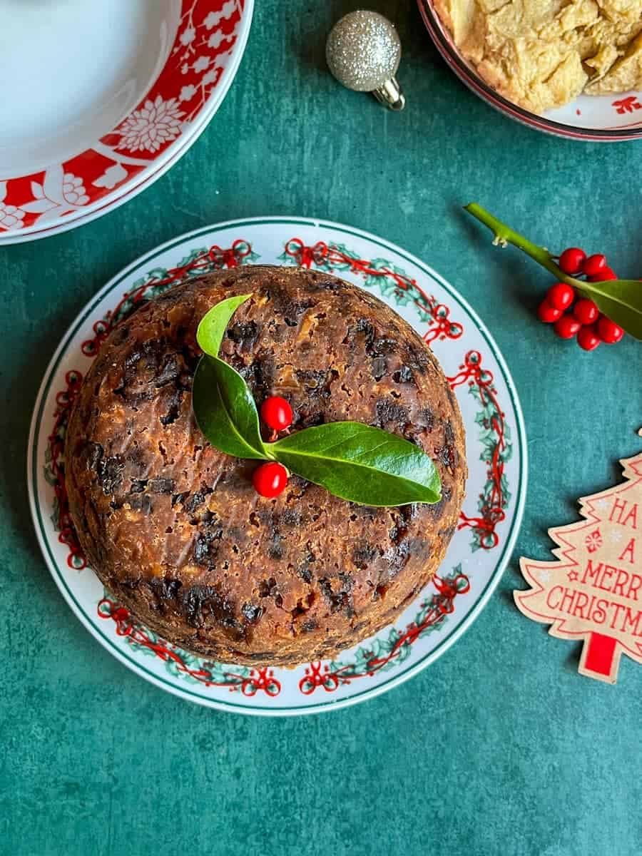 A Christmas pudding topped with a sprig of holly on a red and green Christmas plate. Two red and white bowls, a bowl of brandy butter, Christmas decorations and holly berries can be partially seen.