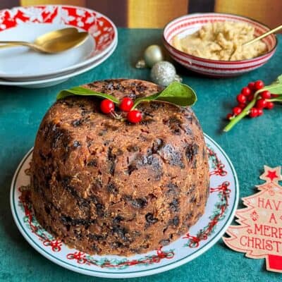 A Christmas pudding with a sprig of holly placed on top on a Christmas plate, a red and white dish of brandy butter with a gold spoon, two red and white bowls with gold spoons and holly berries.