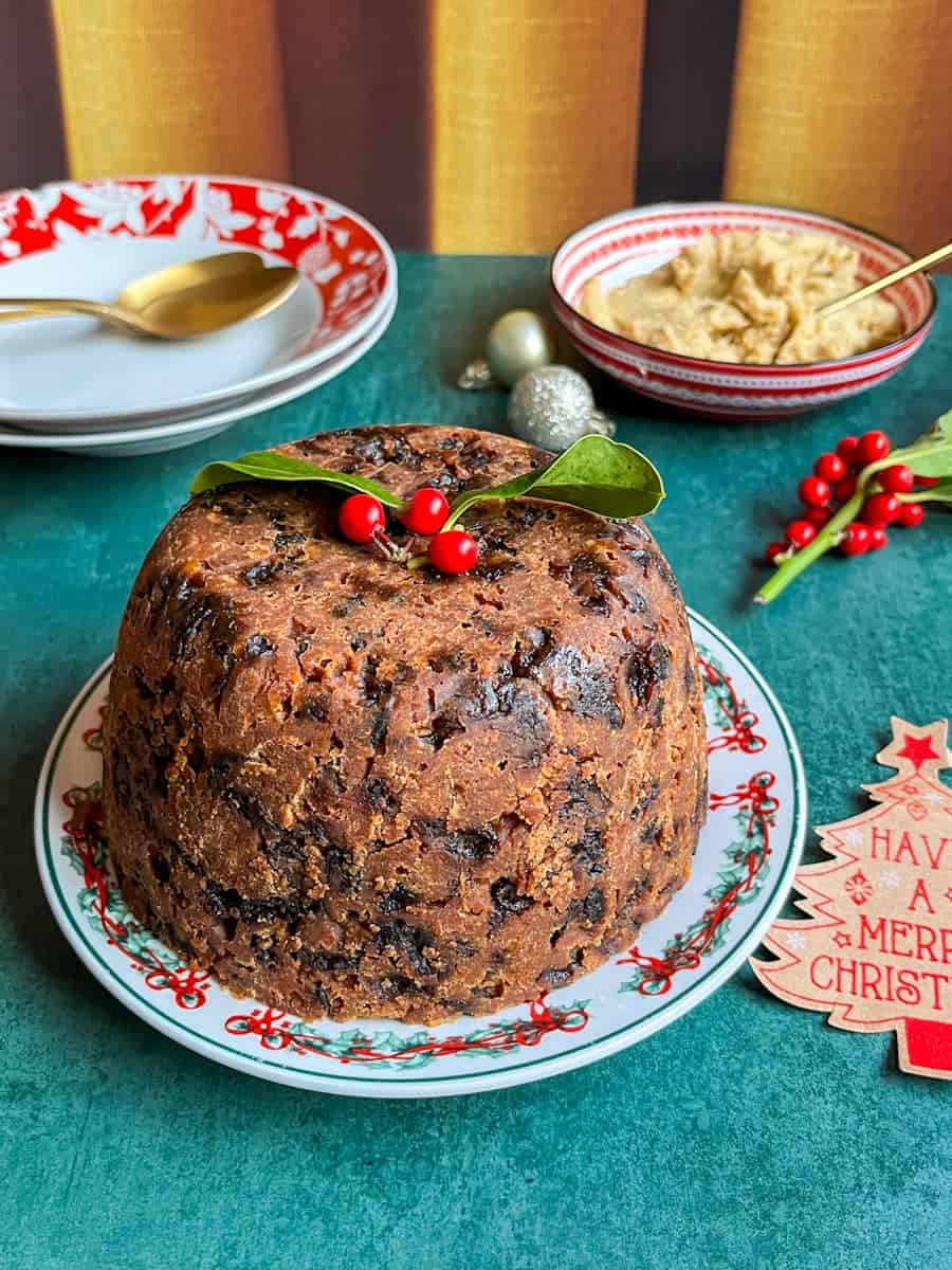 A Christmas pudding with a sprig of holly placed on top on a Christmas plate, a red and white dish of brandy butter with a gold spoon, two red and white bowls with gold spoons and holly berries.