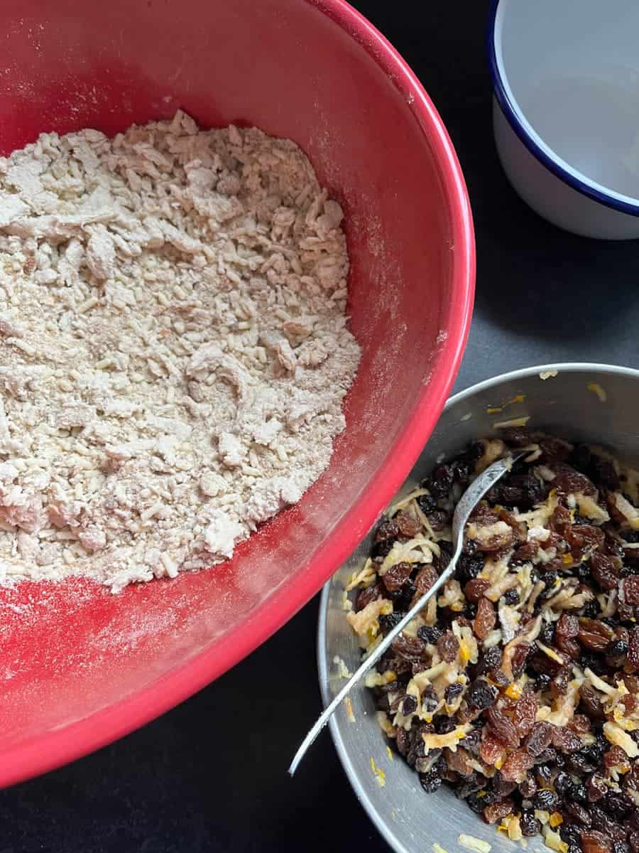 A large red bowl of flour and suet with grated butter and a silver bowl of dried fruit and grated apple.
