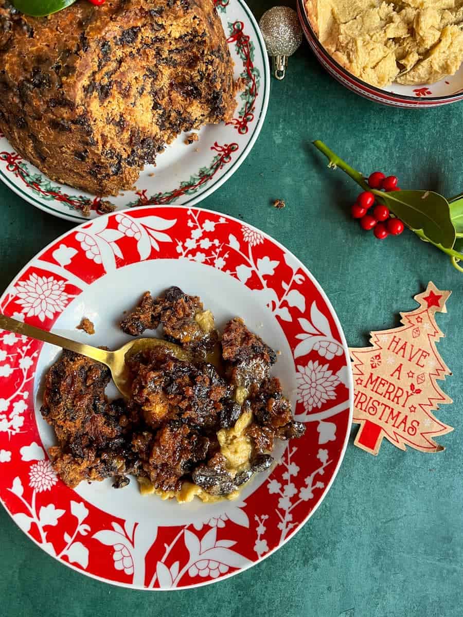 A red and white bowl of Christmas pudding with brandy butter, a Christmas pudding on a red and green festive plate, a bowl of brandy butter and a sprig of holly.