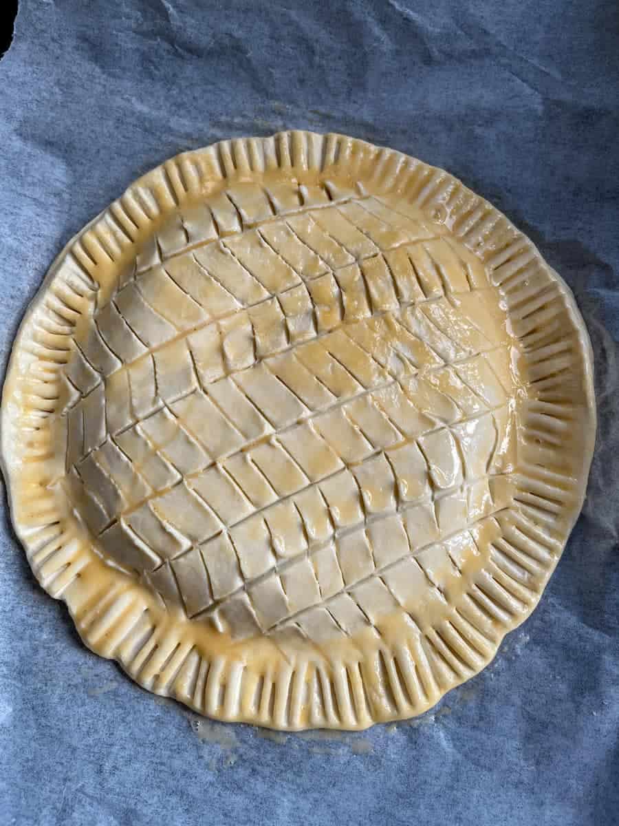 An unbaked Galette de Rois scored in a decorative pattern and glazed with egg wash on a lined baking tray.