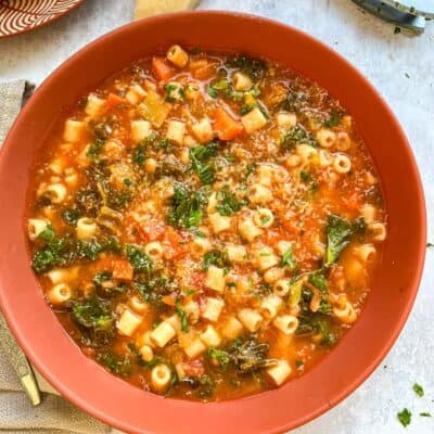 An orange bowl of minestrone soup with chopped carrots, pasta, beans and kale.