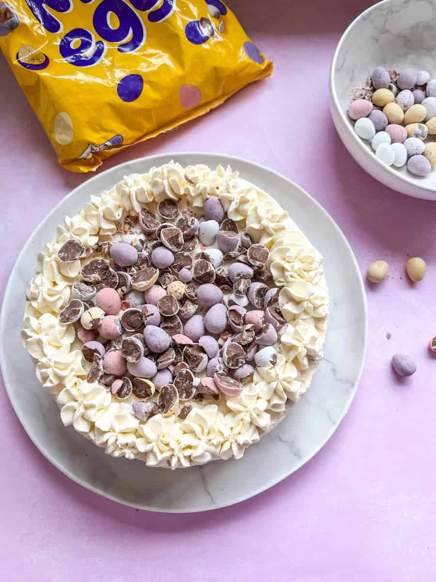 A mini egg cheesecake decorated with whipped cream and crushed mini eggs on a white plate and a white bowl and large packet of mini eggs.