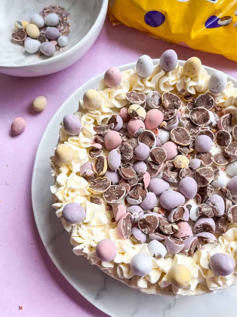A no bake cheesecake topped with whipped cream and crushed mini eggs on a white plate.