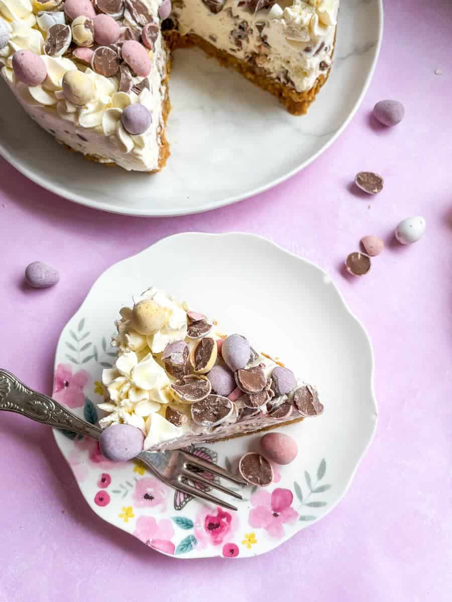 A slice of mini egg cheesecake with a silver fork on a white plate decorated with butterflies and flowers and a large mini egg cheesecake on a white plate.