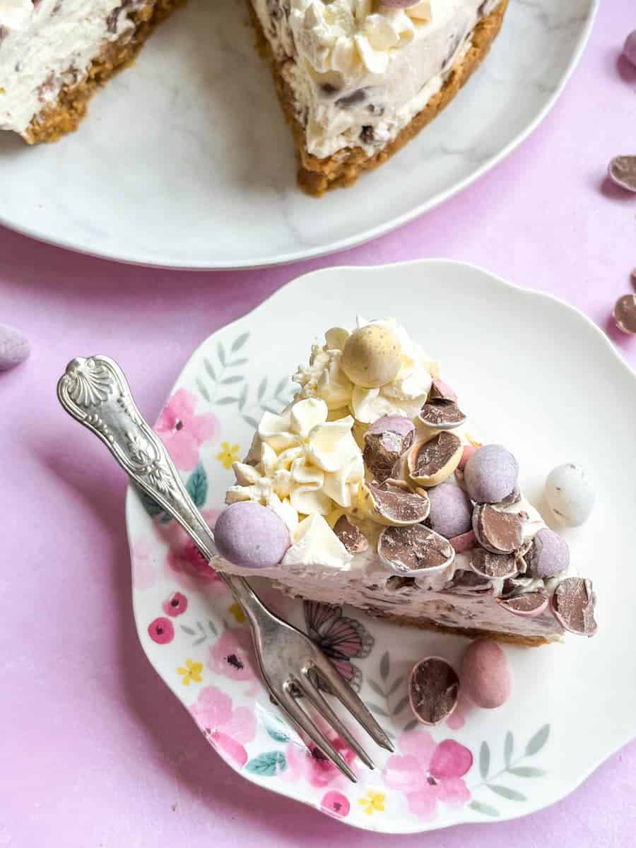 A slice of no bake cheesecake topped with mini eggs with whipped cream on a floral plate with a silver fork.