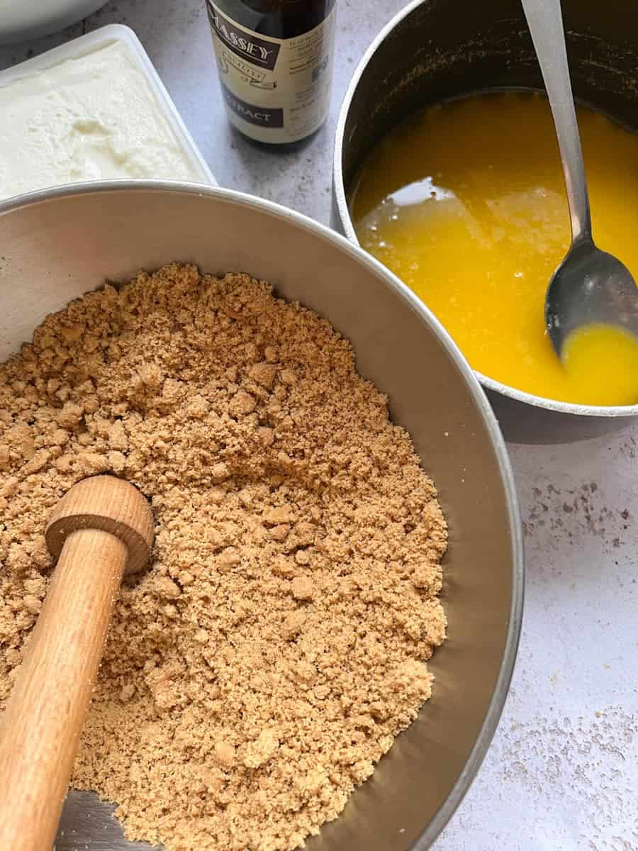 A silver bowl of crushed biscuits, a small pan of melted butter with a silver spoon, a bottle of vanilla extract and a tub of cream cheese.