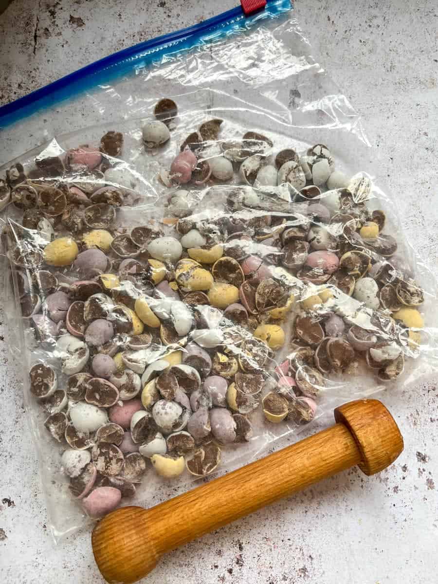 Crushed mini chocolate eggs inside a plastic bag and a small wooden rolling pin.