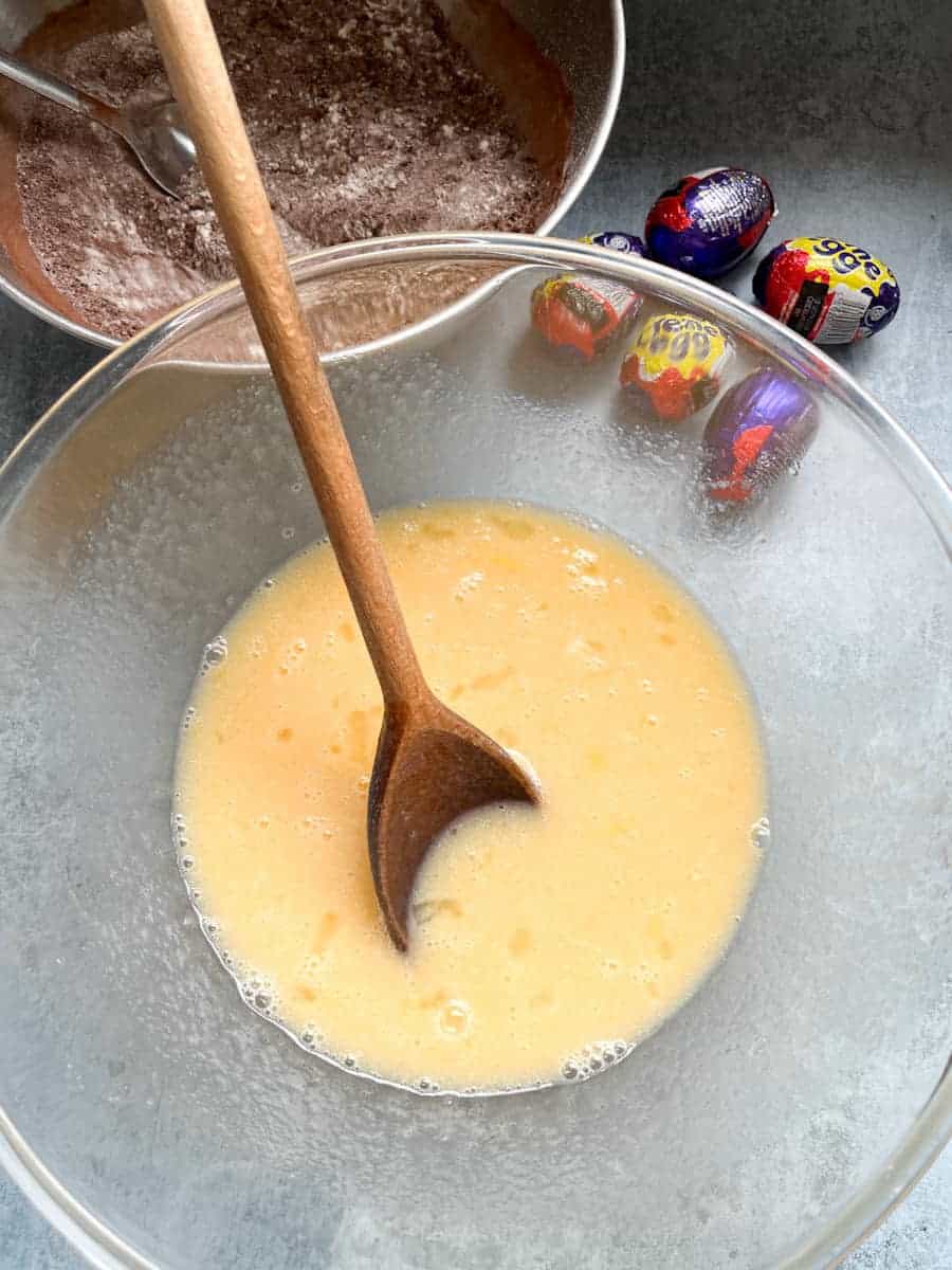 A glass mixing bowl of beaten eggs with a wooden spoon, a silver bowl of flour and cocoa powder and five creme eggs.