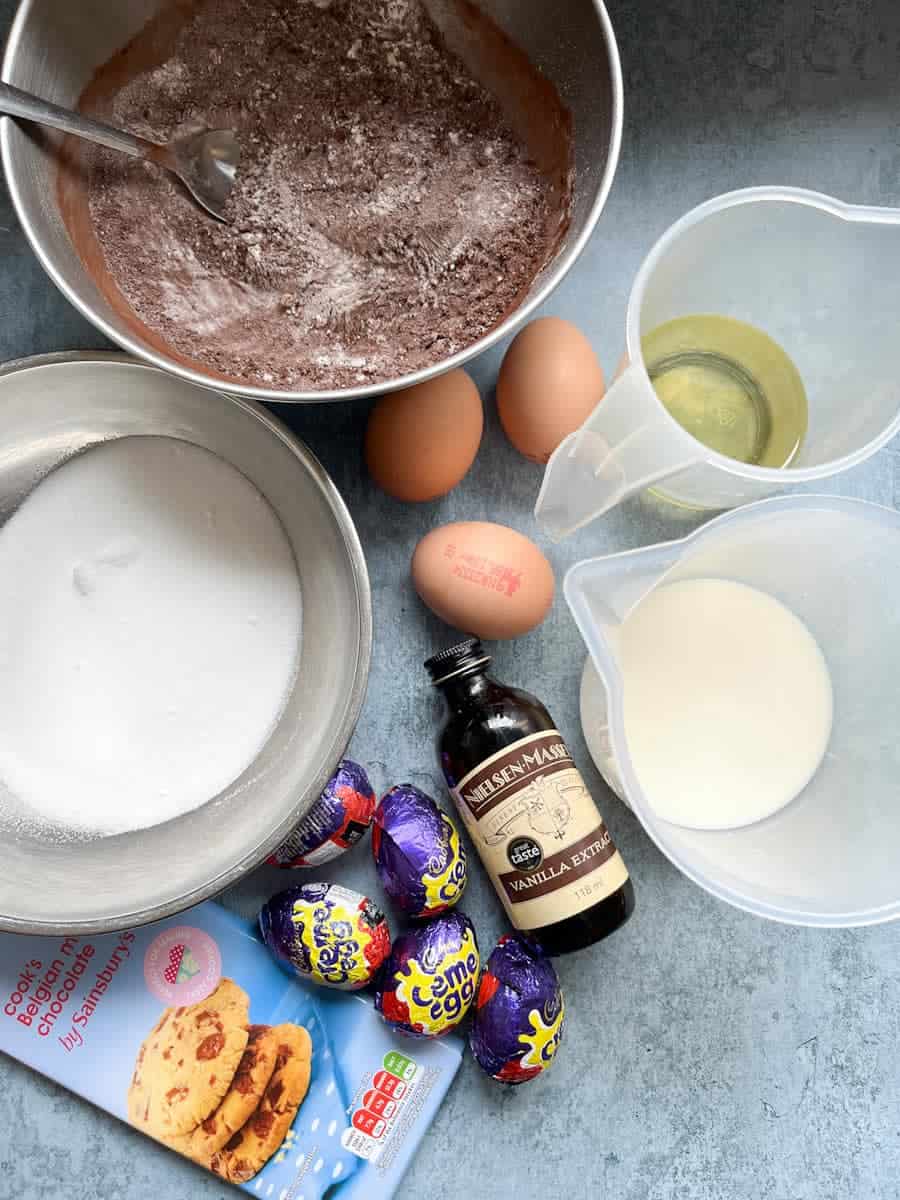 A bowl of flour and cocoa powder, a bowl of white sugar, a jug of oil, a jug of milk, three eggs, a bottle of vanilla extract, five creme eggs and a bar of chocolate.