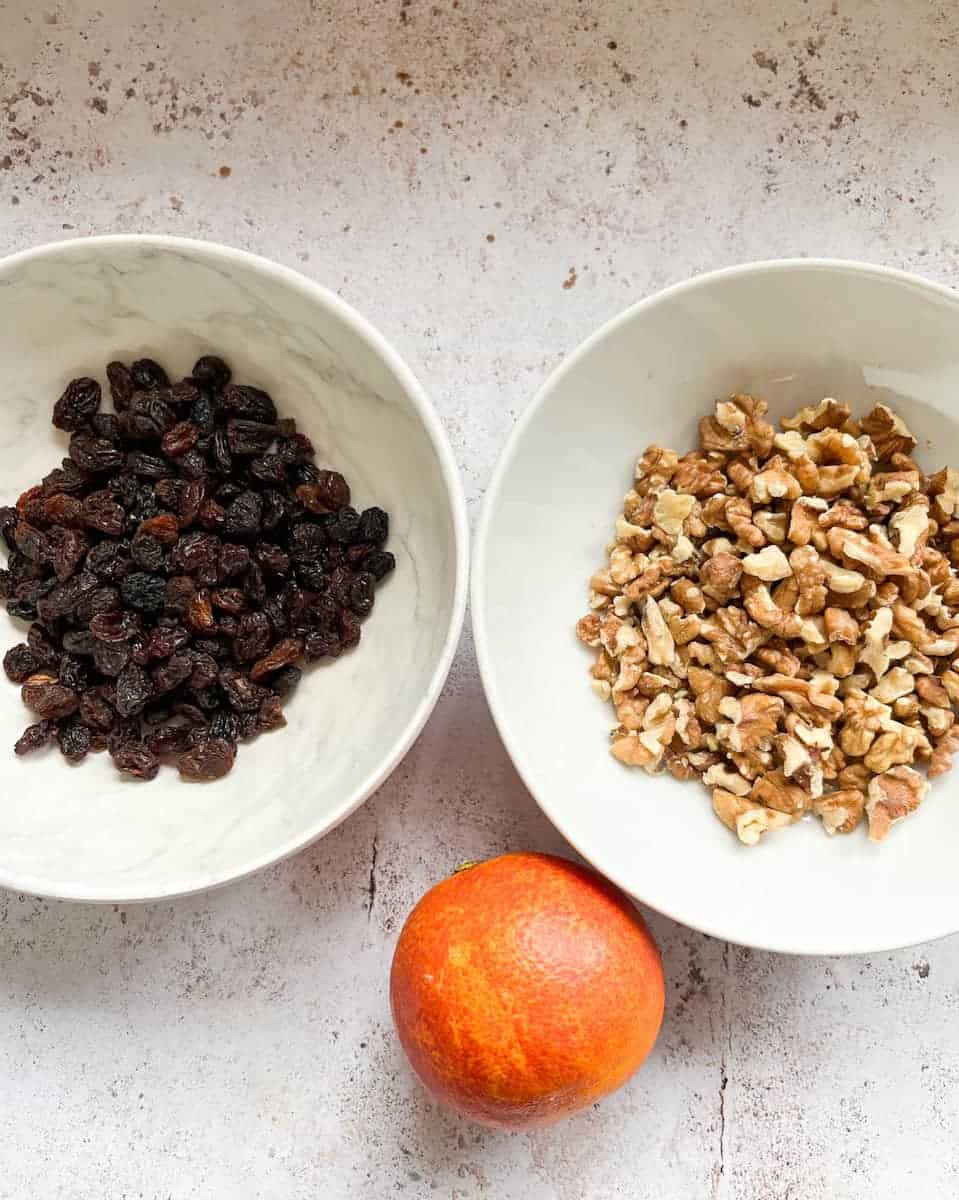 A white bowl of raisins, a white bowl of walnuts and an orange on a grey and white surface. 